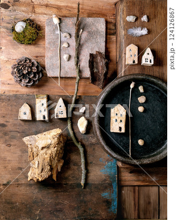 Flat lay creative design rustic composition with miniature ceramic houses, pebbles, shell, moss, pine cone, feather, twigs on wooden background. Natural materials arrangement. Flat lay creative design rustic composition with miniature ceramic houses, pebbles, shell, moss, pine cone, feather, twigs on wooden background. Natural materials arrangement. 124126867