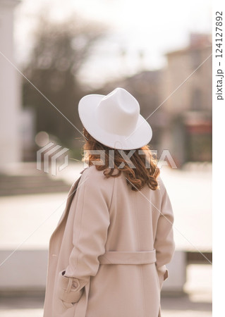 Back view of happy woman wearing hat and coat walking down street on sunny spring day. People, lifestyle, travel and vacations concept 124127892