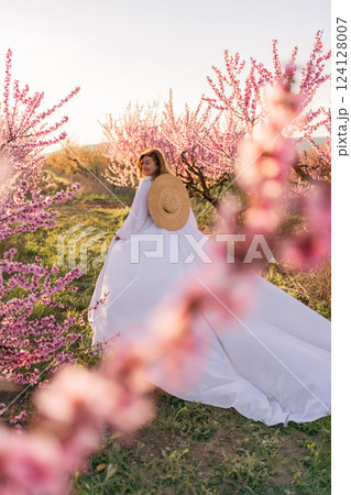 Woman blooming peach orchard. Against the backdrop of a picturesque peach orchard, a woman in a long white dress and hat enjoys a peaceful walk in the park, surrounded by the beauty of nature. 124128007