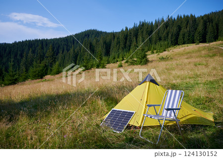 Yellow tent and striped folding chair set up in grassy field, with solar panel nearby. Scene surrounded by lush green trees and rolling hills under clear blue sky, suggesting peaceful camping spot. Yellow tent and striped folding chair set up in grassy field, with solar panel nearby. Scene surrounded by lush green trees and rolling hills under clear blue sky, suggesting peaceful camping spot. 124130152