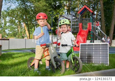 Two boys wearing helmets give thumbs-up while standing with balance bikes next to solar panel on grass. Behind them, playground with red slides and climbing structures surrounded by trees and fence. 124130171