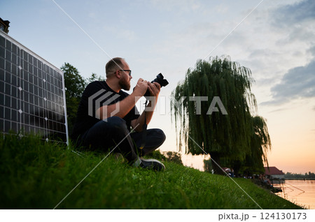 Photographer sits on grass at sunset, capturing serene landscape with camera. Solar panel set up beside, emphasizing use of renewable energy to power equipment. Sustainable practices in photography. 124130173