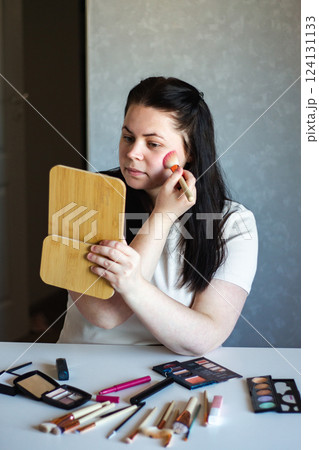 Vertical image of Millennial Caucasian woman doing self make up applying blush on cheeks with make up brush while looking in mirror. Getting ready. Beauty concept. 124131133