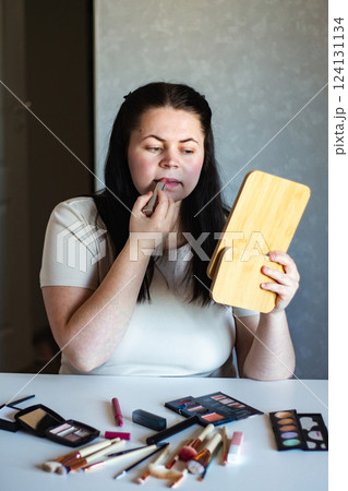 Vertical image of Millennial Caucasian woman doing self make up applying lipstick to lips while looking in mirror. Getting ready. Beauty concept. 124131134