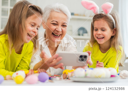 Happy Easter. Two girls sisters twins grandmother painting eggs watching video on phone enjoying time together. Happy family preparing for Easter. Good time at home. Christian festival tradition Happy Easter. Two girls sisters twins grandmother painting eggs watching video on phone enjoying time together. Happy family preparing for Easter. Good time at home. Christian festival tradition 124132986