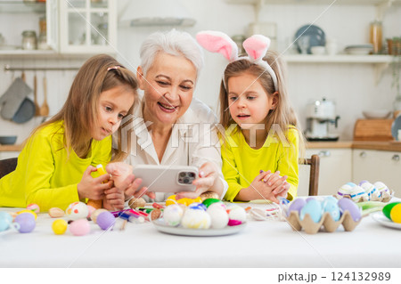 Happy Easter. Two girls sisters twins grandmother painting eggs watching video on phone enjoying time together. Happy family preparing for Easter. Good time at home. Christian festival tradition Happy Easter. Two girls sisters twins grandmother painting eggs watching video on phone enjoying time together. Happy family preparing for Easter. Good time at home. Christian festival tradition 124132989