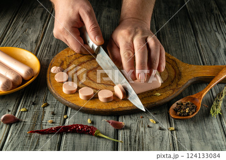 Hands are seen cutting sausage into even slices on a wooden board. Spices like black pepper and red chili adorn the workspace, adding a cozy kitchen atmosphere 124133084