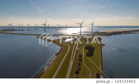 A scenic aerial view of a coastal wind farm with multiple wind turbines standing over the water. Curved roads lead through the landscape, with cars visible. The sun reflects off the water. A scenic aerial view of a coastal wind farm with multiple wind turbines standing over the water. Curved roads lead through the landscape, with cars visible. The sun reflects off the water. 124133453
