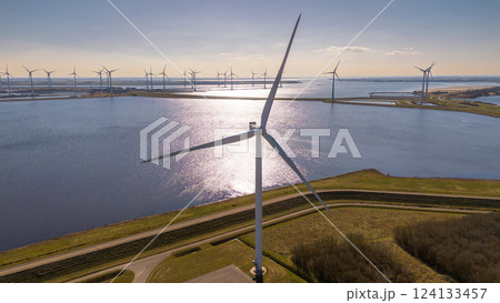 A large wind turbine stands near a coastal waterway, reflecting sunlight. More turbines line the horizon, generating renewable energy. Curved roads and green fields complete this scenic. A large wind turbine stands near a coastal waterway, reflecting sunlight. More turbines line the horizon, generating renewable energy. Curved roads and green fields complete this scenic. 124133457