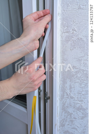 A man's hands insulates windows and balcony doors with foam adhesive tape. Close up. Budget renovation in the apartment. Preparation for the cold snap. Heating problem in apartment buildings 124133797