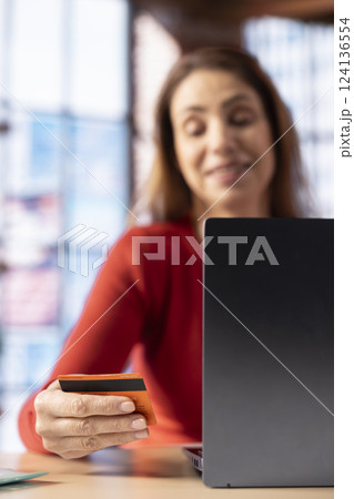Adult holds her card to check balance and oversee budget planning, uses laptop at home office to track her expenses responsibly and confirm payments. Mature woman manages her finances. Adult holds her card to check balance and oversee budget planning, uses laptop at home office to track her expenses responsibly and confirm payments. Mature woman manages her finances. 124136554