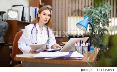 Young woman internist studies the patient's outpatient card, typing the treatment appointment on the computer in the office 124139694