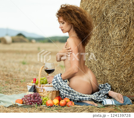 Topless woman in gray checkered unbuttoned shirt holds glass of wine, sitting next to straw pile Topless woman in gray checkered unbuttoned shirt holds glass of wine, sitting next to straw pile 124139728