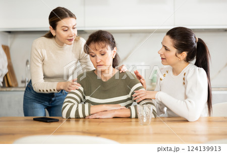 Sad woman sits silently in kitchen, adult girls sympathize and comfort her 124139913