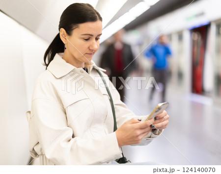 Girl with phone is sitting on metro platform station, scrolling though internet 124140070