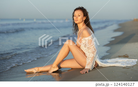 Woman in white lingerie and mesh cape sitting in rays of setting sun on seashore 124140127