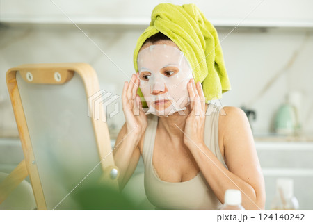 Middle-aged woman applying tissue mask on her facial skin sitting in front of the mirror Middle-aged woman applying tissue mask on her facial skin sitting in front of the mirror 124140242