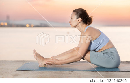 Near sea during sunset, girl performs Pashchimottanasana 124140243