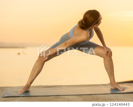 Near sea during sunset, girl performs variations of Himavanasana 124140443