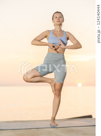 Sedulous woman doing warrior one leg pose of yoga on mat on beach at sunrise Sedulous woman doing warrior one leg pose of yoga on mat on beach at sunrise 124140444