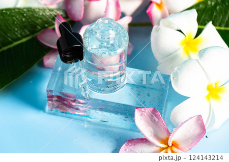 Clear serum in a dropper bottle rests on a blue surface with vibrant frangipani flowers and leaves. 124143214