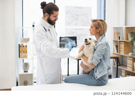 Veterinarian with beard and woman holding white dog in bright clinic discussing x-ray results. Male veterinarian wearing coat and gloves, woman with short hair sitting, cheerful dog in her arms. 124144600