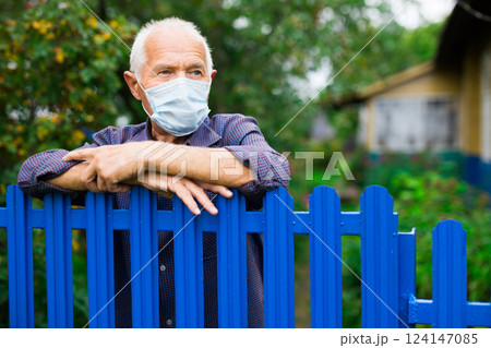 Portrait of elderly man in protective mask at fence of his country house in the village. Isolation concept during pandemic Portrait of elderly man in protective mask at fence of his country house in the village. Isolation concept during pandemic 124147085