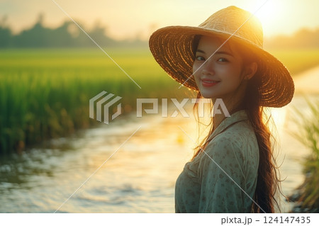 a woman managing an irrigation system in a rice field, standing on a levee with water flowing nearby, wearing traditional farming attire and smiling under bright sunlight 124147435