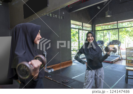Arab middle eastern muslim woman working out while looking in the mirror and lifting a dumbbell at the gym. Fitness exercise and healthy lifestyle concept 124147486