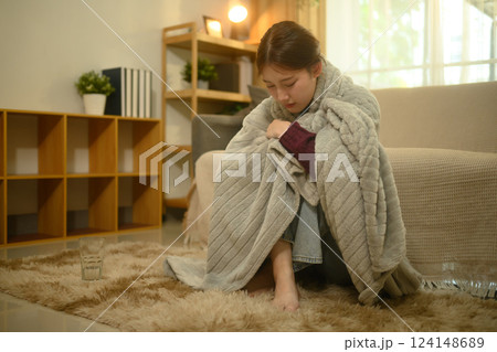 Sick woman sitting on the floor wrapped in a gray blanket. Illness and home recovery concept 124148689