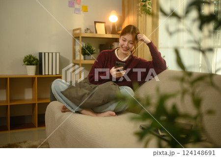 Smiling young asian woman enjoying social media on smartphone sitting on couch at home 124148691