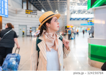 Young Caucasian elegant pretty woman in straw hat, wearing backpack, holds her passport to check in for a flight. Waiting in airport Young Caucasian elegant pretty woman in straw hat, wearing backpack, holds her passport to check in for a flight. Waiting in airport 124150784
