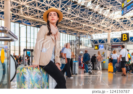 Young Caucasian woman, elegantly dressed, is sitting on a suitcase at the airport or train station. There are people in the background out of focus. The concept of travel and vacation 124150787