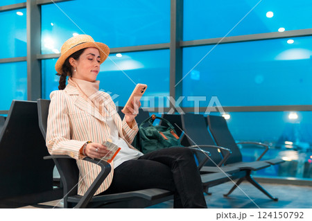 Mid shot of young elegant Caucasian woman in a hat and jacket using smartphone in airport waiting area sitting in chair. Concept of boarding at terminal before flight Mid shot of young elegant Caucasian woman in a hat and jacket using smartphone in airport waiting area sitting in chair. Concept of boarding at terminal before flight 124150792