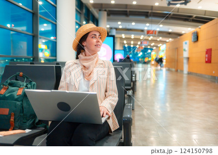 Young Caucasian elegant happy woman is sitting on a chair in waiting room of airport and working on a laptop. Copy space. Concept of remote work and travel Young Caucasian elegant happy woman is sitting on a chair in waiting room of airport and working on a laptop. Copy space. Concept of remote work and travel 124150798