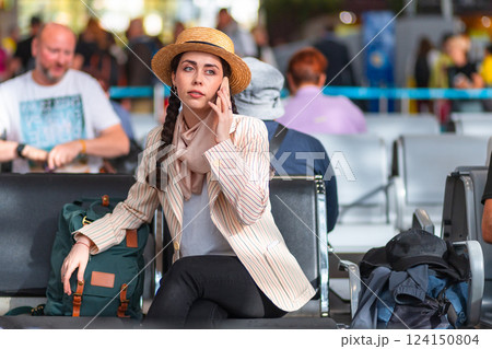 A young Caucasian woman in a hat and jacket is sitting in an armchair in the waiting room and talking on a smartphone. Waiting to board the plane A young Caucasian woman in a hat and jacket is sitting in an armchair in the waiting room and talking on a smartphone. Waiting to board the plane 124150804