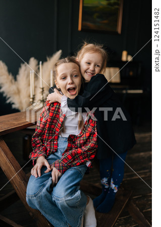 Playful young sisters embracing, making silly faces, sharing genuine laughter in warm home interior, revealing close sibling bond and joyful connection, vertical portrait, smiling looking away. Playful young sisters embracing, making silly faces, sharing genuine laughter in warm home interior, revealing close sibling bond and joyful connection, vertical portrait, smiling looking away. 124151482