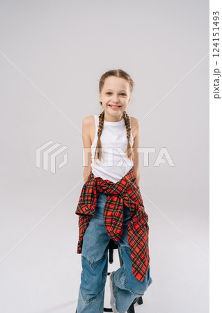 Studio vertical portrait of joyful 10 years old girl with braided hair posing smiling looking at camera. Pretty preteen child wearing white tank top, blue jeans, red checkered shirt knotted at waist. Studio vertical portrait of joyful 10 years old girl with braided hair posing smiling looking at camera. Pretty preteen child wearing white tank top, blue jeans, red checkered shirt knotted at waist. 124151493