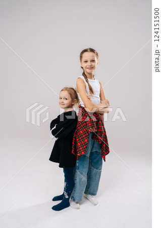 Full length vertical portrait of two young sisters posing back to back in studio setting, arms crossed and radiating confidence, showcasing strong bond filled with love and happiness looking at camera Full length vertical portrait of two young sisters posing back to back in studio setting, arms crossed and radiating confidence, showcasing strong bond filled with love and happiness looking at camera 124151500