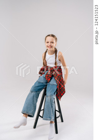 Full length vertical portrait of joyful preteen girl with braids, wearing jeans and plaid shirt tied around waist, sitting on stool, laughing heartily on white isolated background, looking at camera 124151523
