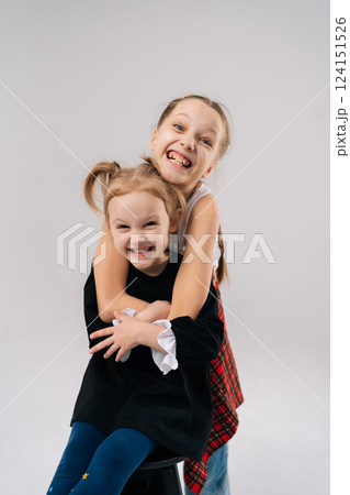 Vertical portrait of two young sisters embracing and laughing, creating heartwarming portrait of sibling love and happiness in professional studio setting with white background, looking at camera. Vertical portrait of two young sisters embracing and laughing, creating heartwarming portrait of sibling love and happiness in professional studio setting with white background, looking at camera. 124151526