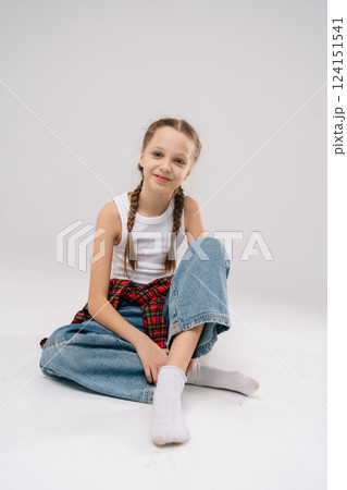 Vertical portrait of pretty young girl with braided hair wearing white tank top and red plaid shirt, sitting cross-legged with cheerful expression against neutral backdrop, smiling looking at camera, 124151541