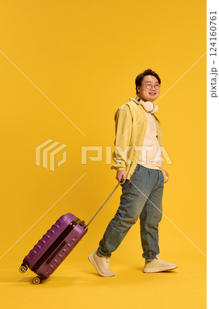 Cheerful smiling young Asian man walking, dressed casual attire and headphones, with luggage, ready to trip against sunny yellow background. 124160761