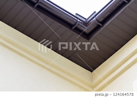 Close-up detail of cottage house corner with brown metal planks siding and roof with steel gutter rain system. Roofing, construction, drainage pipes installation and connection concept. 124164578