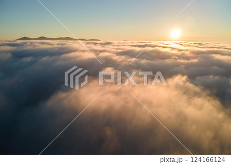 Aerial view of colorful sunrise over white dense fog with distant dark silhouettes of mountain hills on horizon Aerial view of colorful sunrise over white dense fog with distant dark silhouettes of mountain hills on horizon 124166124