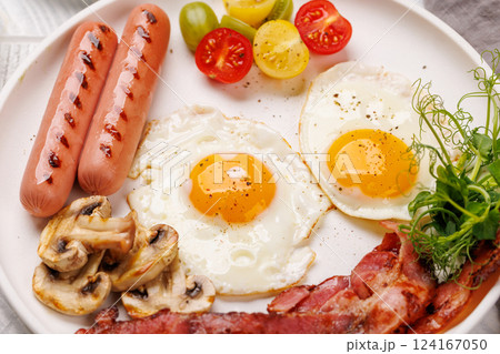 A classic breakfast plate featuring sunny-side-up eggs, crispy bacon, sausages, and fresh tomatoes 124167050