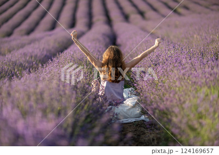 A woman is sitting in a field of lavender flowers. She is stretching her arms out in front of her. 124169657