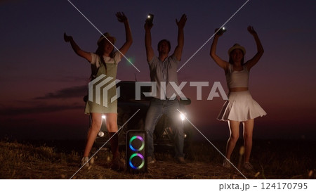 Cheerful company dances by car in evening. People friendly best close friends family on road trip stopped at night jumping partying to music next to glowing rainbow music column with flashlights. 124170795
