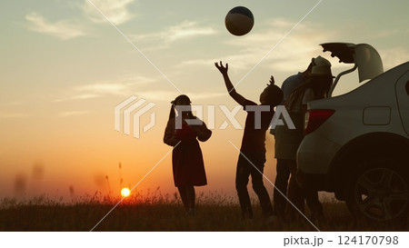 Playful family at sunset by car. cheerful children parents going on travel road trip by automobile packing baggage putting luggage in trunk kids playing ball. Packing preparation for family picnic. 124170798