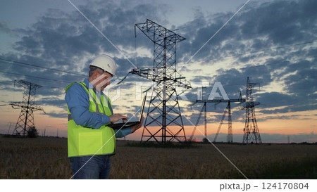 Skilled technician analyses data on laptop at power transmission lines in dusk rural field. Electrician in helmet with laptop controls power generation substation equipment at evening countryside 124170804